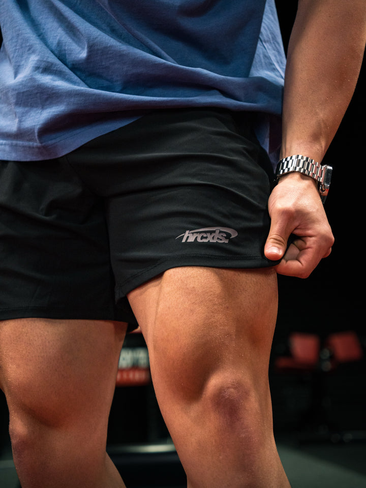 Close-up of an athlete pulling the hem of black HERCXLES shorts in a gym