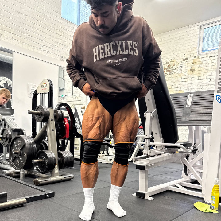 Athlete in a brown HERCXLES Lifting Club hoodie displaying his quadriceps in a gym