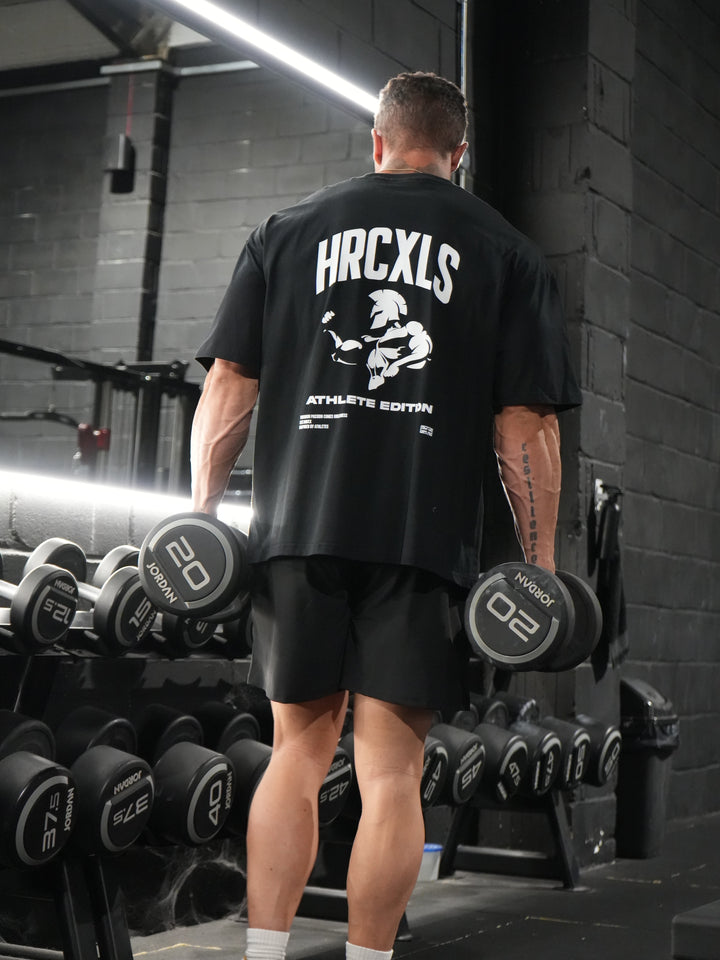 Athlete photographed from behind holding dumbbells in a black HRCXLS Athlete Edition oversized T-shirt in the gym
