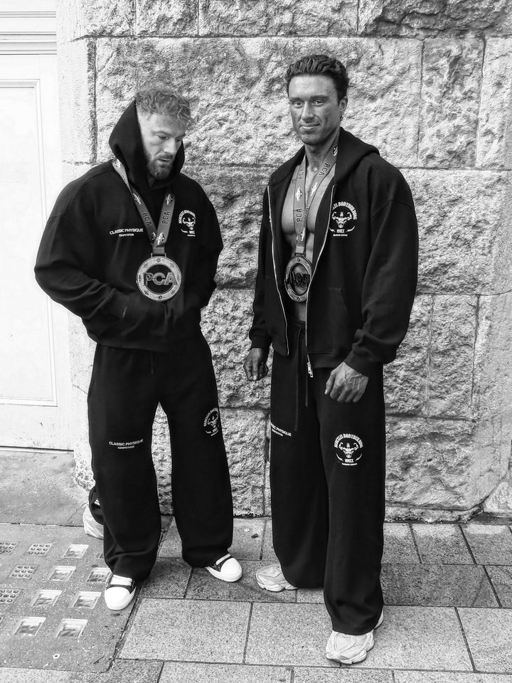 Black-and-white photo of two bodybuilding competitors wearing medals and matching black competition hoodies and sweatpants