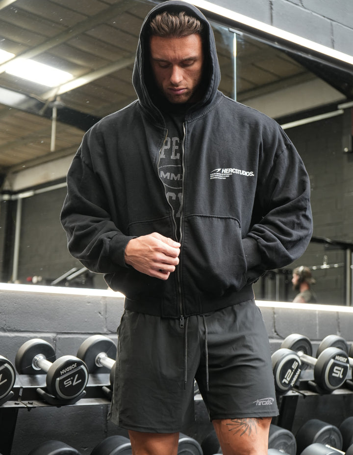 Athlete wearing a black HERCSTUDIOS oversized zip-up hoodie and black shorts in front of a dumbbell rack
