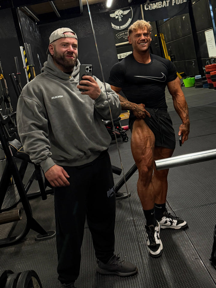 Two bodybuilders pose for a gym mirror selfie; one wears a grey HERCXLES hoodie and backwards cap, while the other wears a black T-shirt and shorts