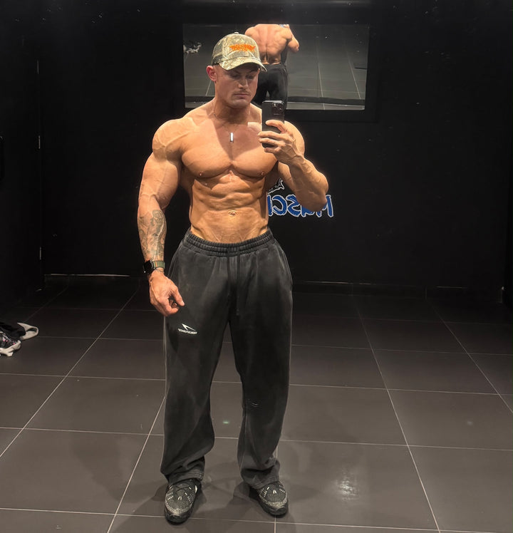 Shirtless bodybuilder in a camouflage cap takes a gym mirror selfie wearing charcoal HERCSTUDIOS fleece sweatpants